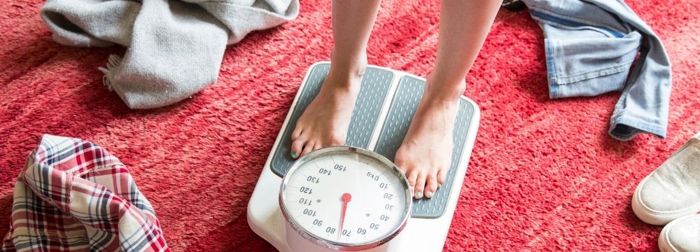 Woman stepping on scale on red rug with clothing scattered around bathroom scale