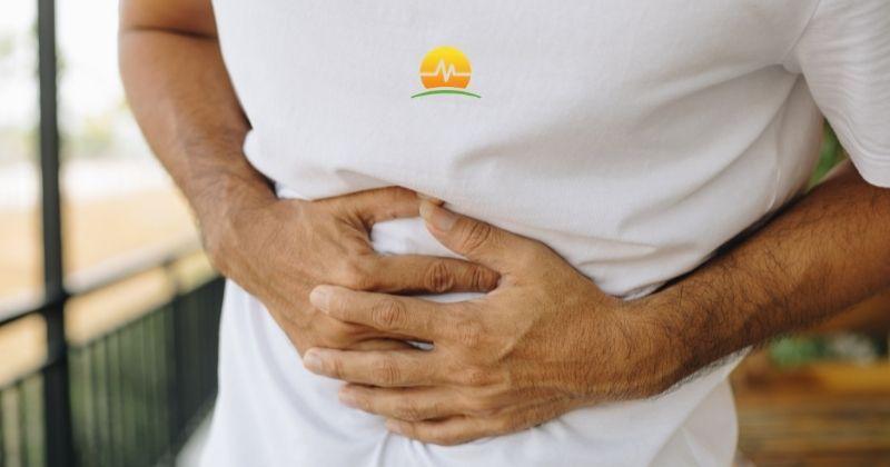 Man in white tee shirt holds pressure on abdomen to support his hernia while he waits for hernia repair surgery. MASJax logo at top center. 