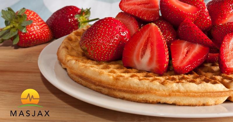 Waffle on white plate topped with sliced fresh strawberries. MasJax logo in the bottom left corner.