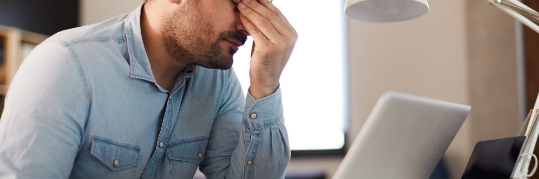 Man sitting at desk with laptop open holding between eyes, stressed out 
