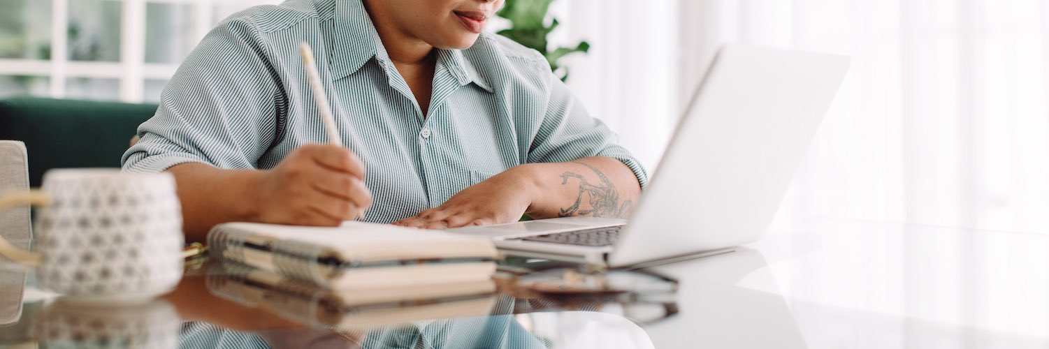 Plus size woman working at office space looking at laptop and writing in notebook 