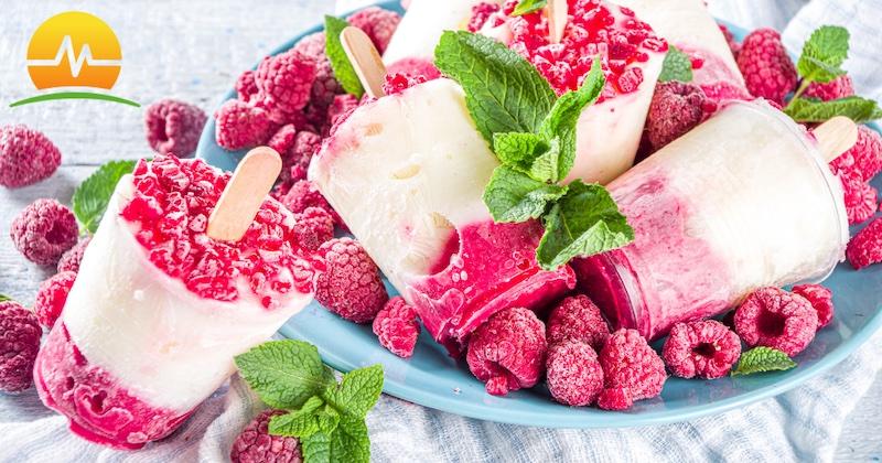 Healthy raspberry vanilla cream popsicles on a blue plate surrounded by fresh raspberries and mint. Memorial Advances Surgery logo in top left corner.